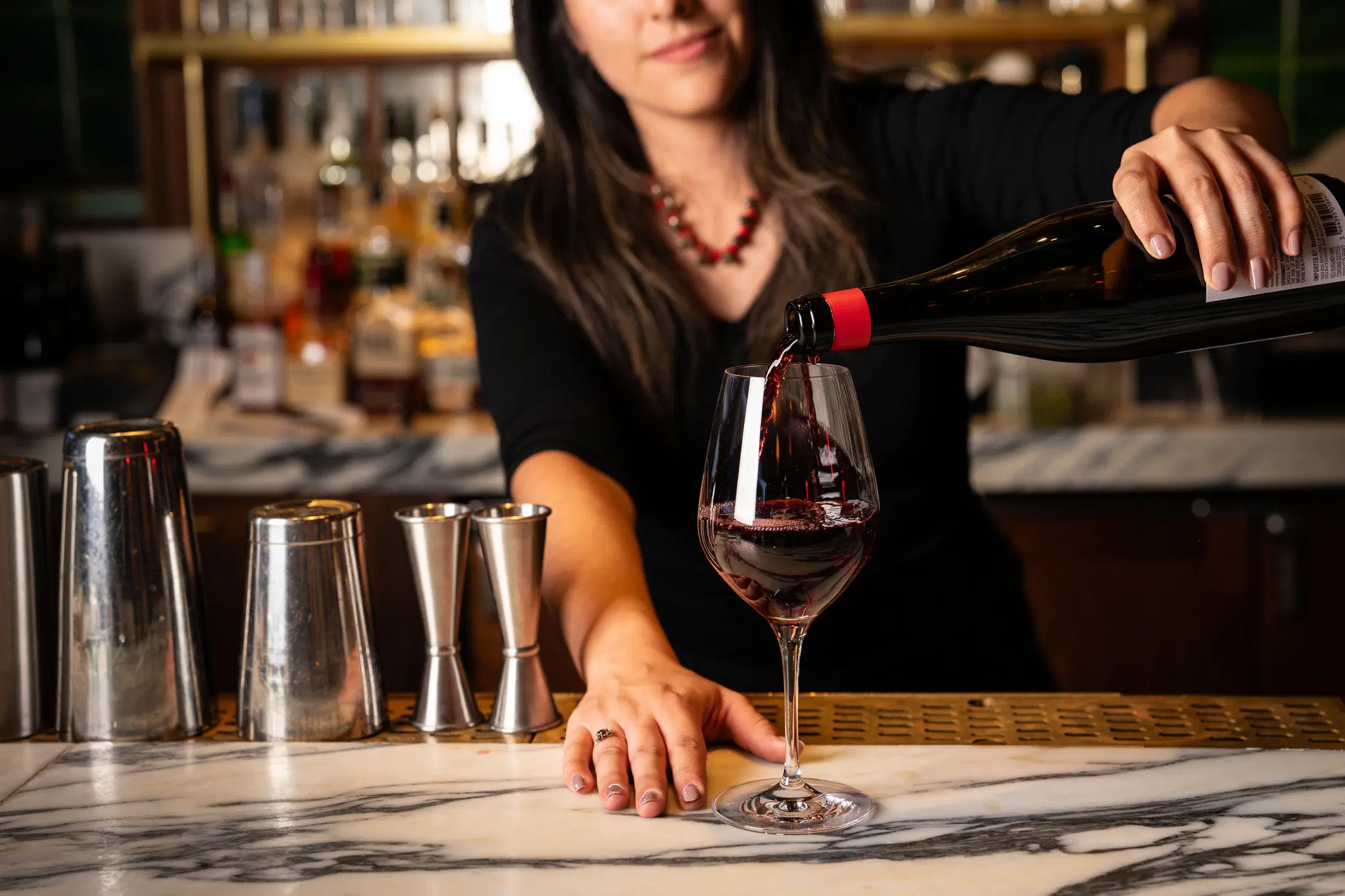 A bartender pours red wine into a glass at a marble bar counter, with cocktail shakers and bottles in the background. 