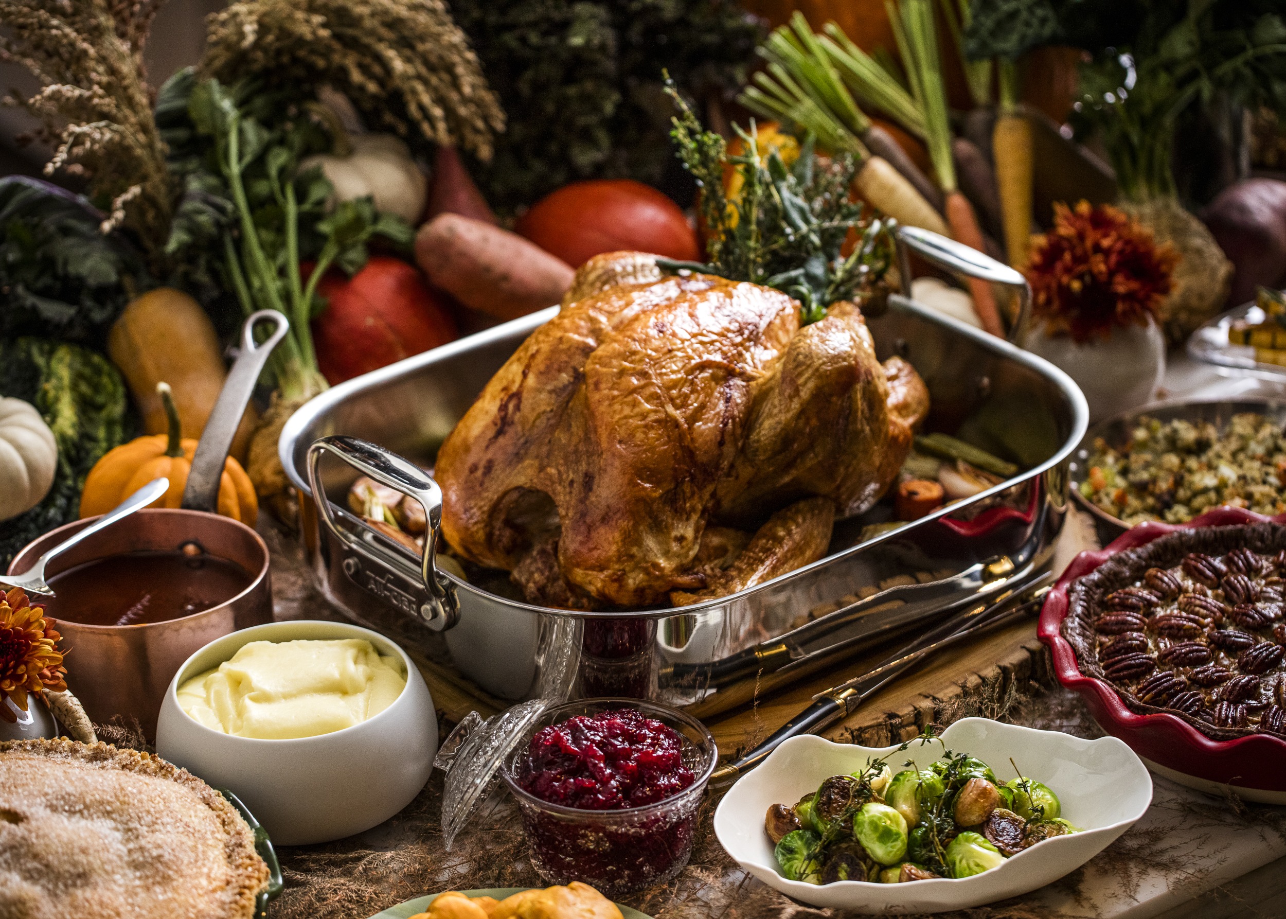 A Thanksgiving feast featuring a roasted turkey in a pan surrounded by dishes such as mashed potatoes, cranberry sauce, Brussels sprouts, stuffing, and a variety of vegetables and pumpkins. 