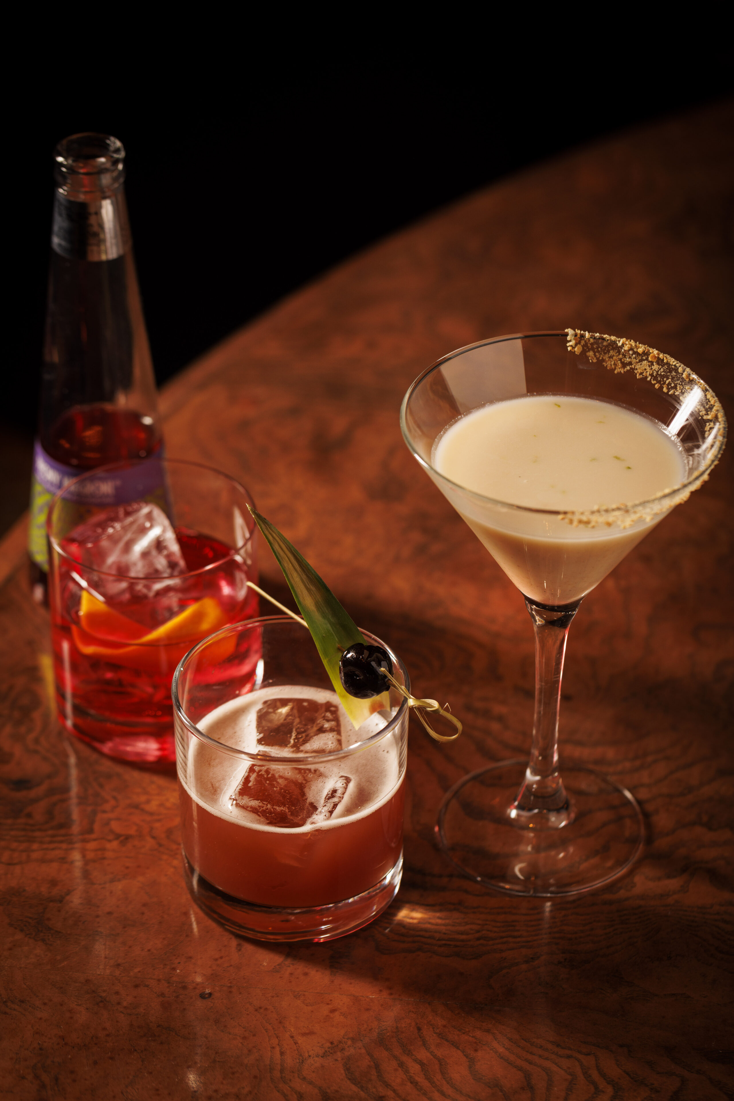 Three mocktails on a wooden table: a red drink with ice and an orange slice beside a bottle, a brown drink with ice and a cherry garnish, and a creamy martini with a sugared rim. 