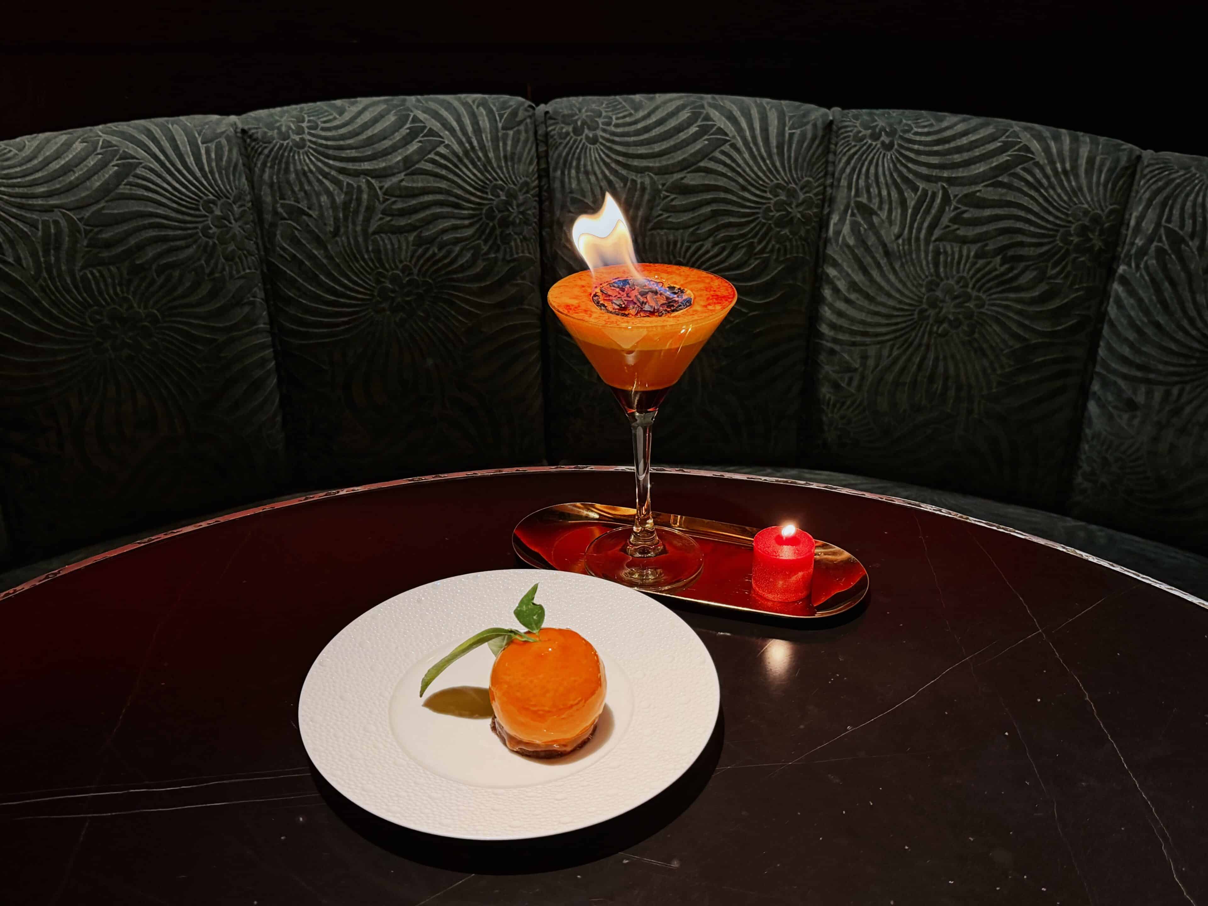 A flaming orange cocktail in a martini glass sits next to a small red candle on a dark round table, with an orange dessert garnished with a mint leaf on a white plate. The background features a dark, textured banquette. This is House of the Red Pearl at Tin Building 